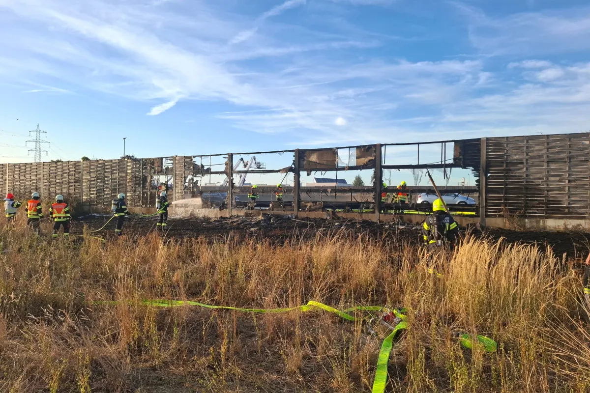 Das Bild zeigt Feuerwehrleute, die eine zerstörte Lärmschutzwand löschen. Im Vordergrund sind brennende Grasflächen sichtbar, während der Himmel klar ist.