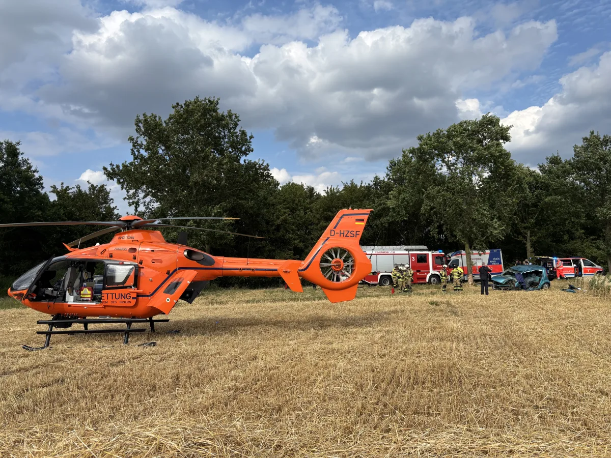 Ein orangefarbener Rettungshubschrauber steht auf einem Feld, umgeben von Feuerwehrfahrzeugen und Einsatzkräften bei einem Autounfall.