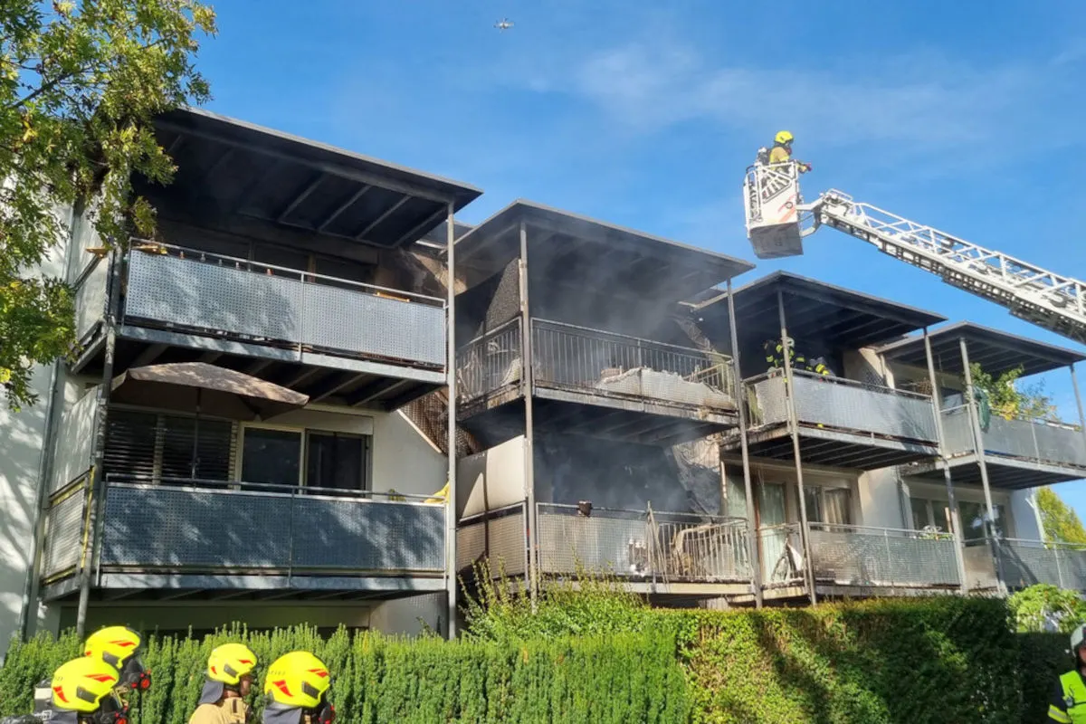 Ein Wohnhaus mit verrauchten Balkonen, Feuerwehrleute sind auf einer Leiter im Einsatz. Rauchsäulen deuten auf ein Feuer hin.