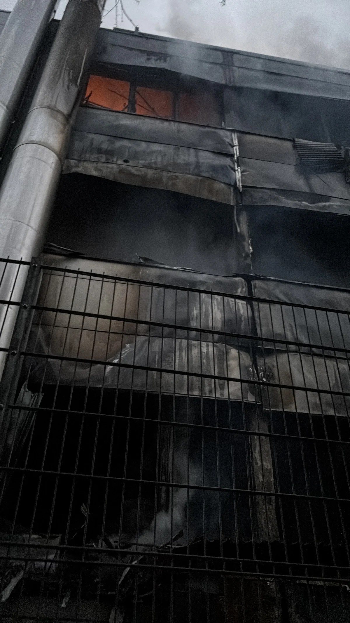 Ein mehrstöckiges Gebäude ist stark beschädigt, mit Rauch und Flammen sichtbar hinter den oberen Fenstern. Der Bereich ist durch einen Metallzaun abgesichert.
