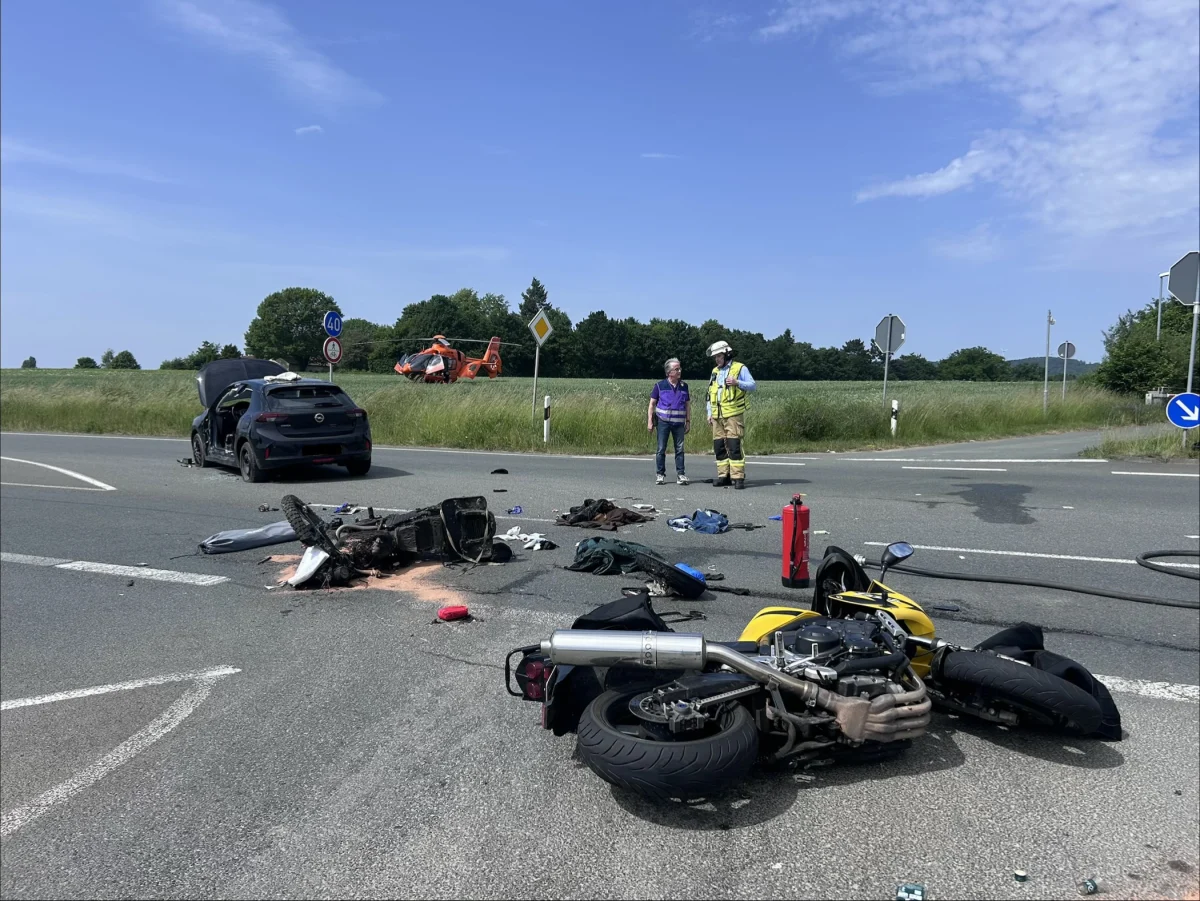 Auf einer Landstraße sind Wrackteile eines Motorrads verstreut, ein beschädigtes Auto steht hinten. Ein Rettungshubschrauber ist im Feld gelandet.