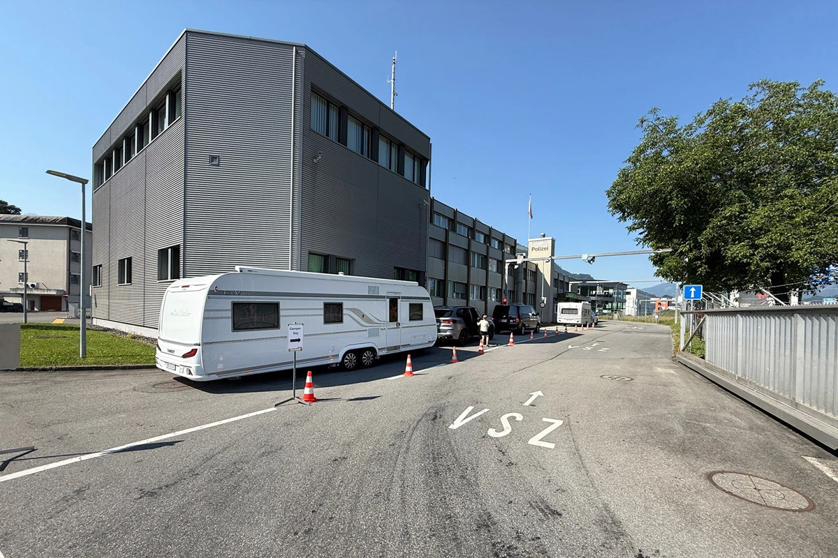 Ein Wohnwagen parkt vor einem großen Polizeigebäude, abgesperrt durch Hütchen. Personen sind in der Nähe sichtbar.