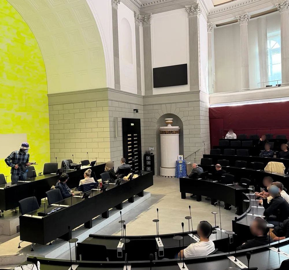 Ein großer Sitzungssaal mit mehreren Personen, die an Schreibtischen sitzen. Die Architektur ist klassisch mit hohen Decken.