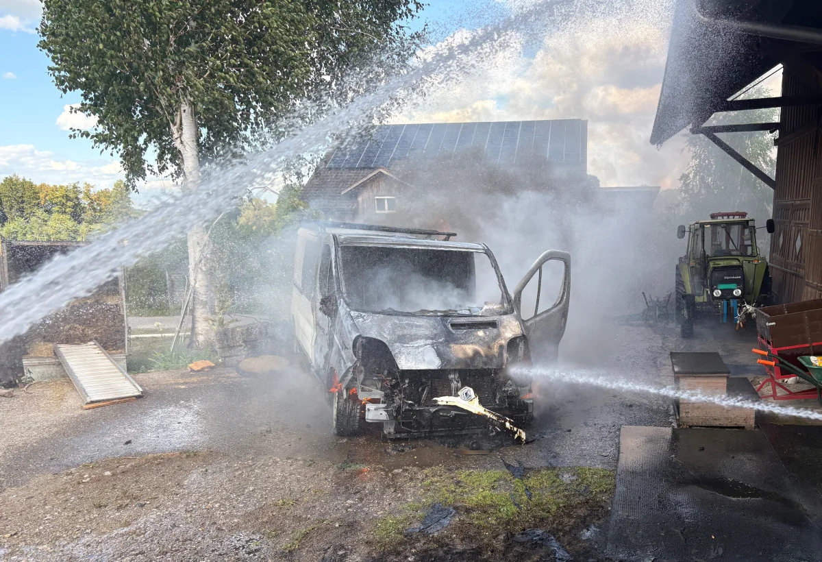 Ein ausgebrannter Transporter in einem ländlichen Bereich wird von der Feuerwehr mit Wasserstrahlen gelöscht. Im Hintergrund ist ein Traktor zu sehen.