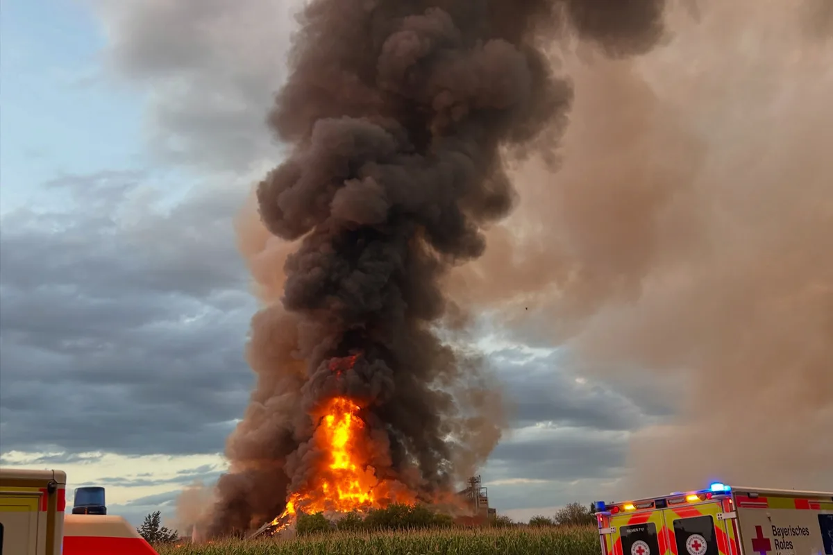 Eine große Feuerstelle erzeugt dichte Rauchwolken, die in den Himmel aufsteigen. Rettungsfahrzeuge sind am Rand der Szene sichtbar.
