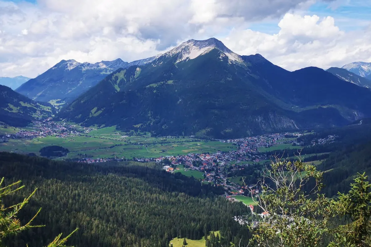 Das Bild zeigt ein weitläufiges Tal, umgeben von hohen Bergen und bewaldeten Flächen unter einem teils bewölkten Himmel.