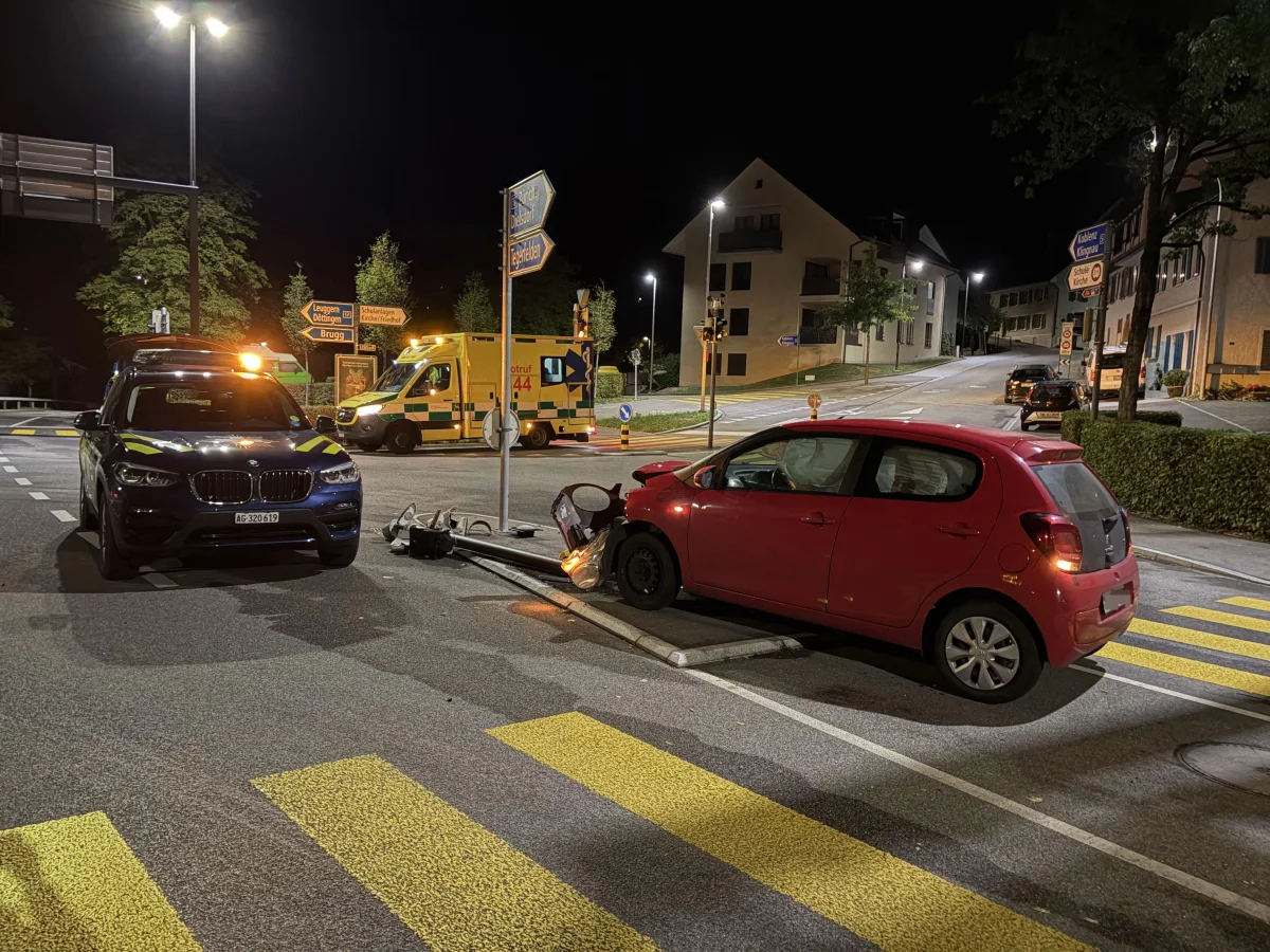 Bei Nacht stößt ein rotes Auto an einer Kreuzung gegen ein Verkehrsschild. Polizei- und Rettungsfahrzeuge sind am Unfallort.