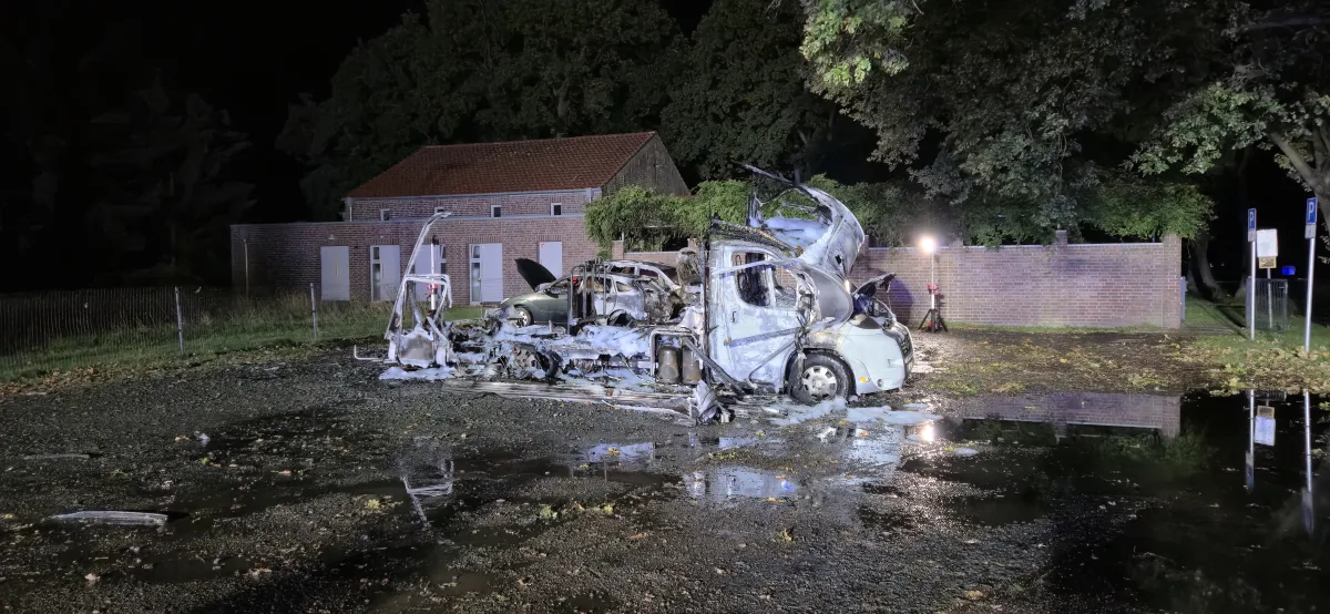 Ein vollkommen ausgebranntes Fahrzeug steht bei Nacht neben einem Backsteingebäude. Der Boden ist feucht und im Hintergrund sind Bäume und ein beleuchtetes Areal zu sehen.