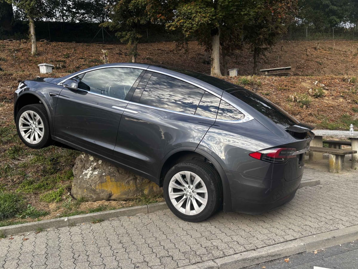 Ein grauer Tesla ist auf einem großen Stein aufgesessen und blockiert den Parkplatz. Die Umgebung ist herbstlich mit Laub auf dem Boden.