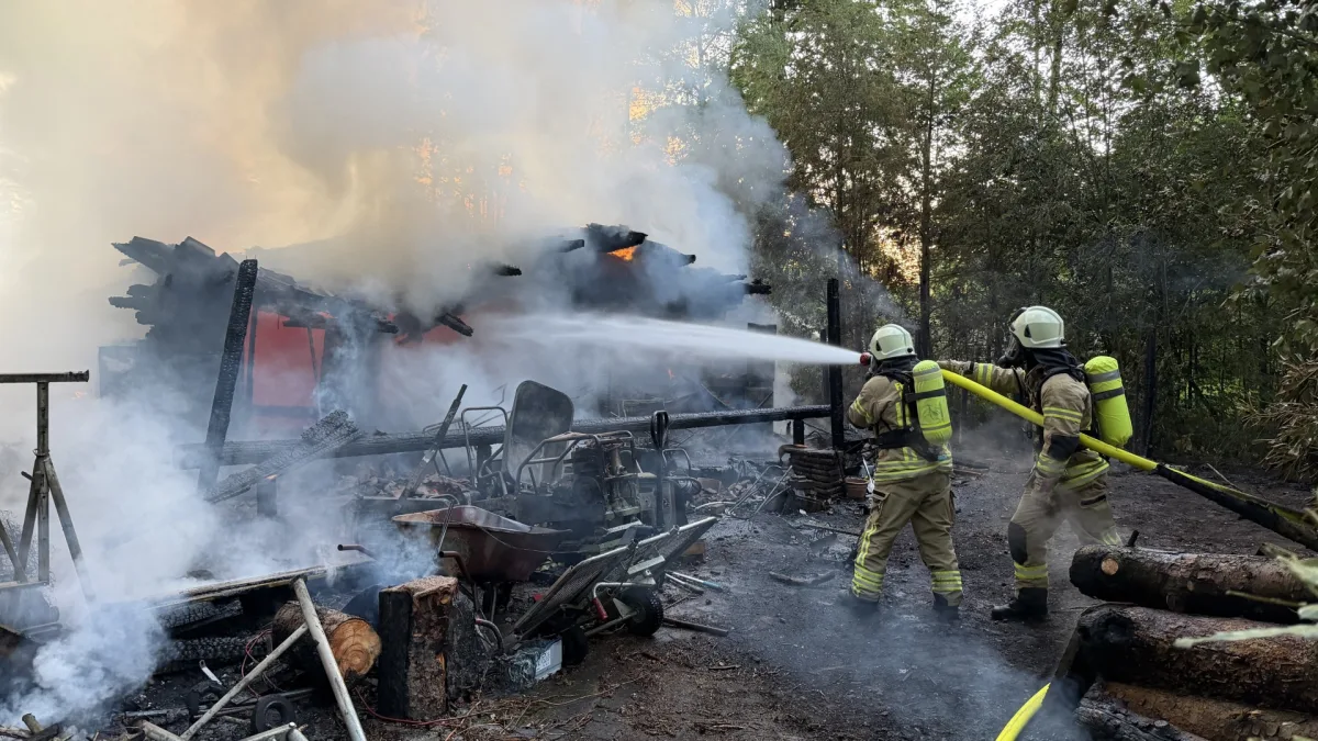 Zwei Feuerwehrleute in Schutzausrüstung löschen ein loderndes Feuer an einem teilweise eingestürzten Gebäude. Rauch und Flammen steigen auf.