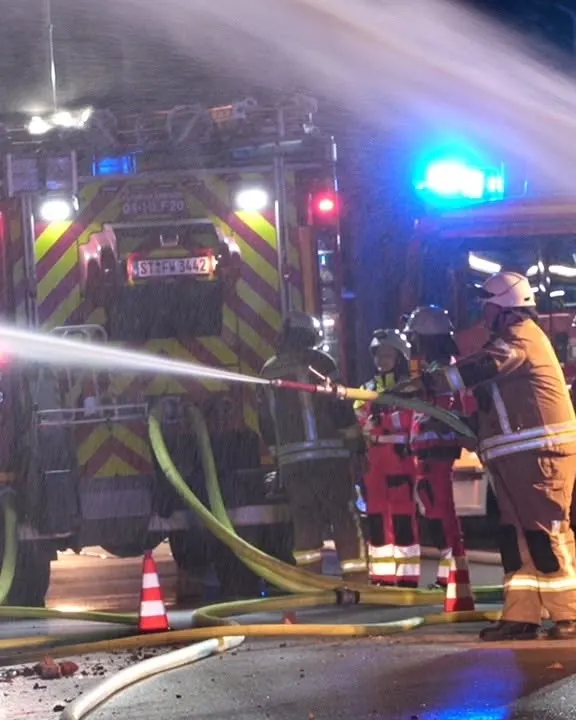 Mehrere Feuerwehrleute bekämpfen nachts ein Feuer mit Wasserschläuchen vor einem Einsatzfahrzeug. Im Hintergrund sind Blaulichter zu sehen.