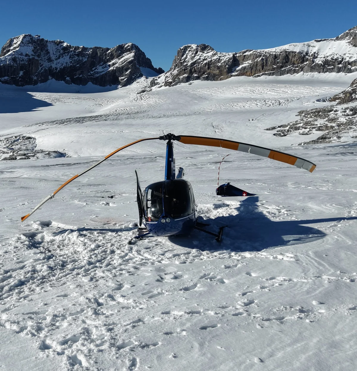 Ein Helikopter liegt mit beschädigtem Rotor auf einem schneebedeckten Gletscher, umgeben von Bergen.