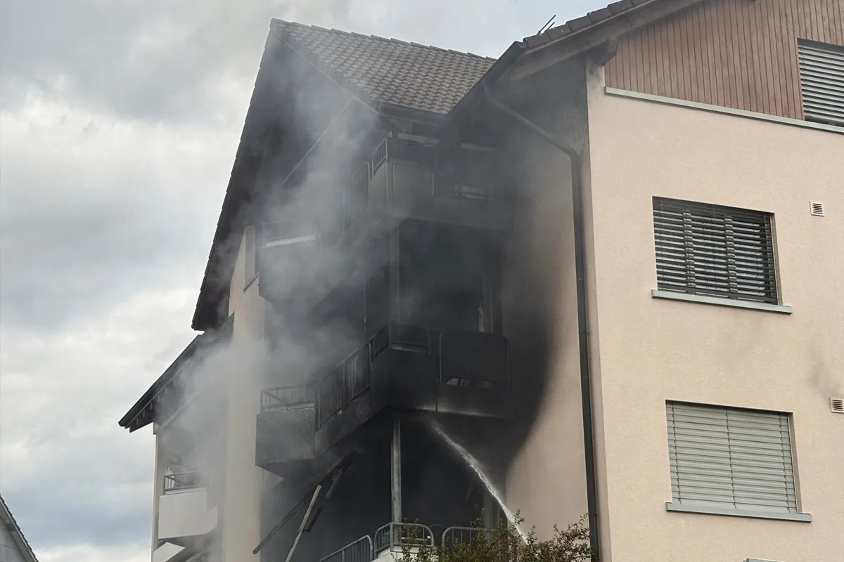 Ein Mehrfamilienhaus mit starkem Rauch und einem Löschschlauch ist abgebildet. Der Brand hat die Fassade schwarz gefärbt.