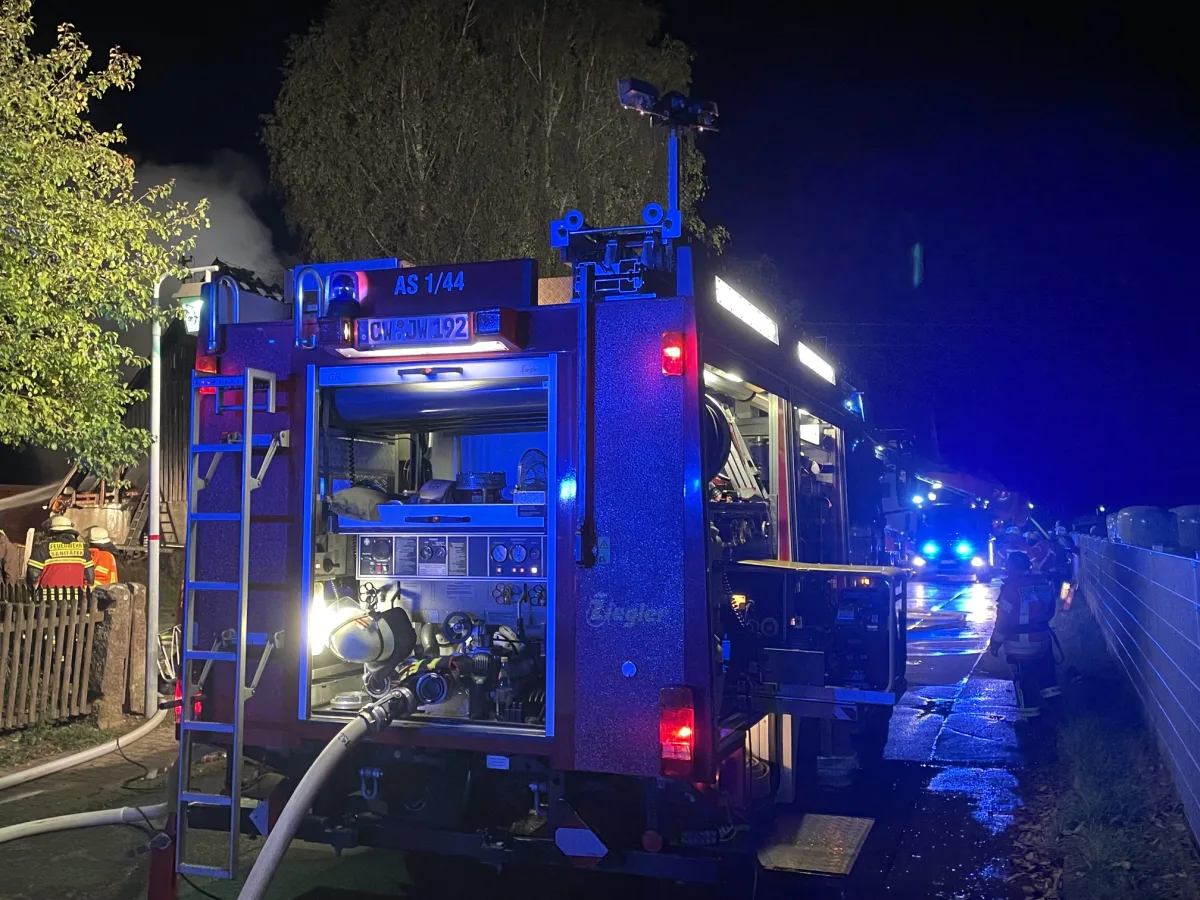 Ein Feuerwehrfahrzeug steht mit geöffnetem Equipmentfach auf einer Straße bei Nacht, Feuerwehrleute sind im Einsatz.