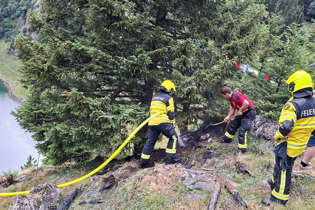 Drei Feuerwehrleute bekämpfen einen kleinen Brand am Waldrand; einer bedient einen Schlauch, während ein anderer mit einer Schaufel arbeitet.