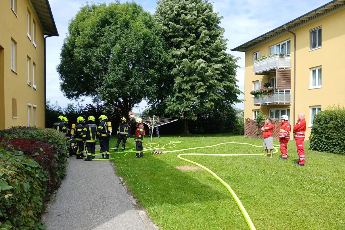 Mehrere Feuerwehrleute in Einsatzkleidung stehen auf einer Wiese vor einem gelben Wohnhaus. Daneben sind weitere Einsatzkräfte in Rot.