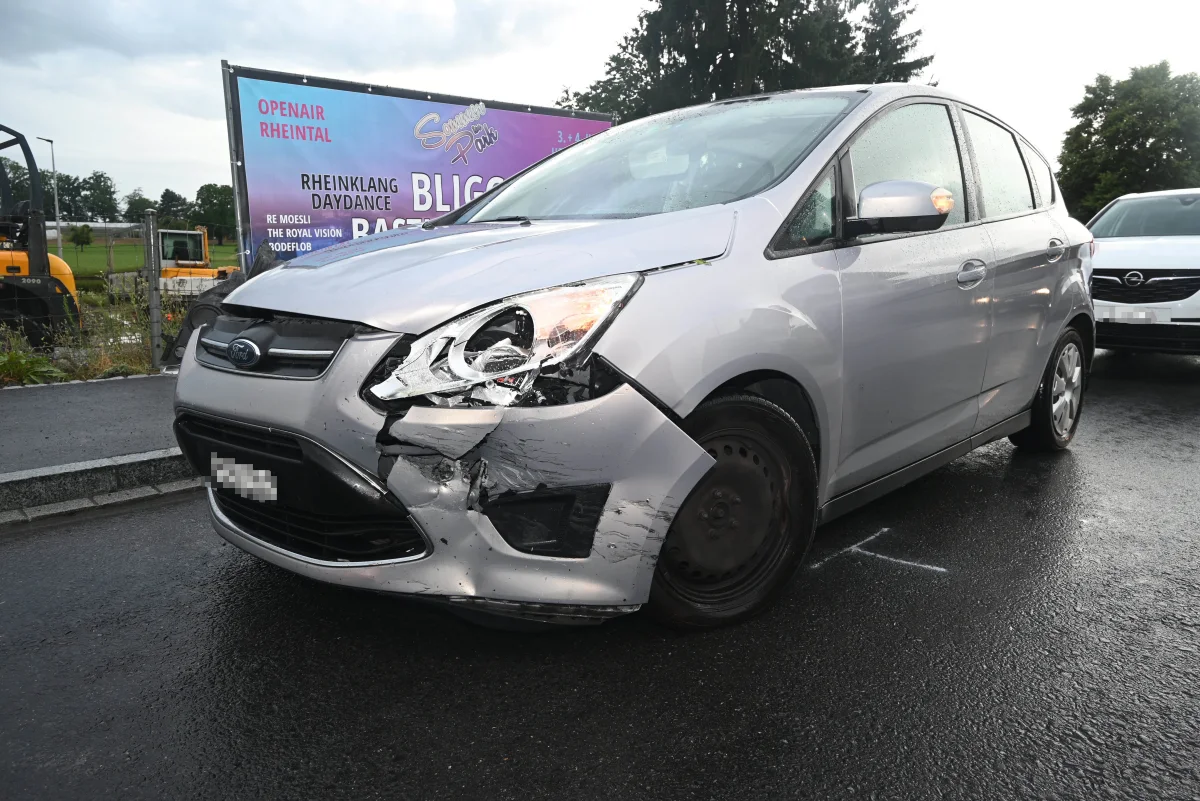 Ein silberner Ford mit beschädigter Front steht auf einer nassen Straße. Im Hintergrund ist ein Werbeplakat sichtbar.