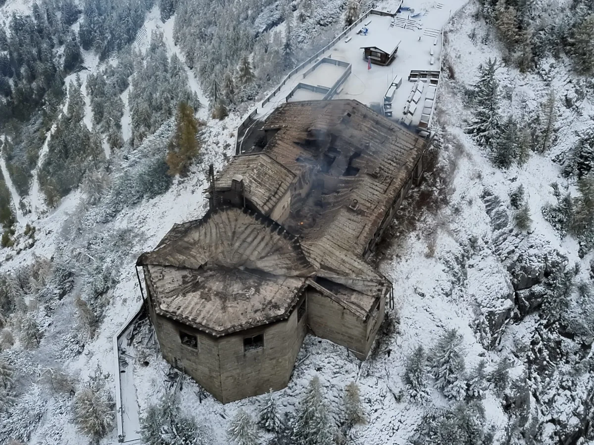 Ein großes, ausgebranntes Gebäude steht in einer verschneiten, bergigen Landschaft. Es ist von Schnee bedeckte Bäume umgeben.