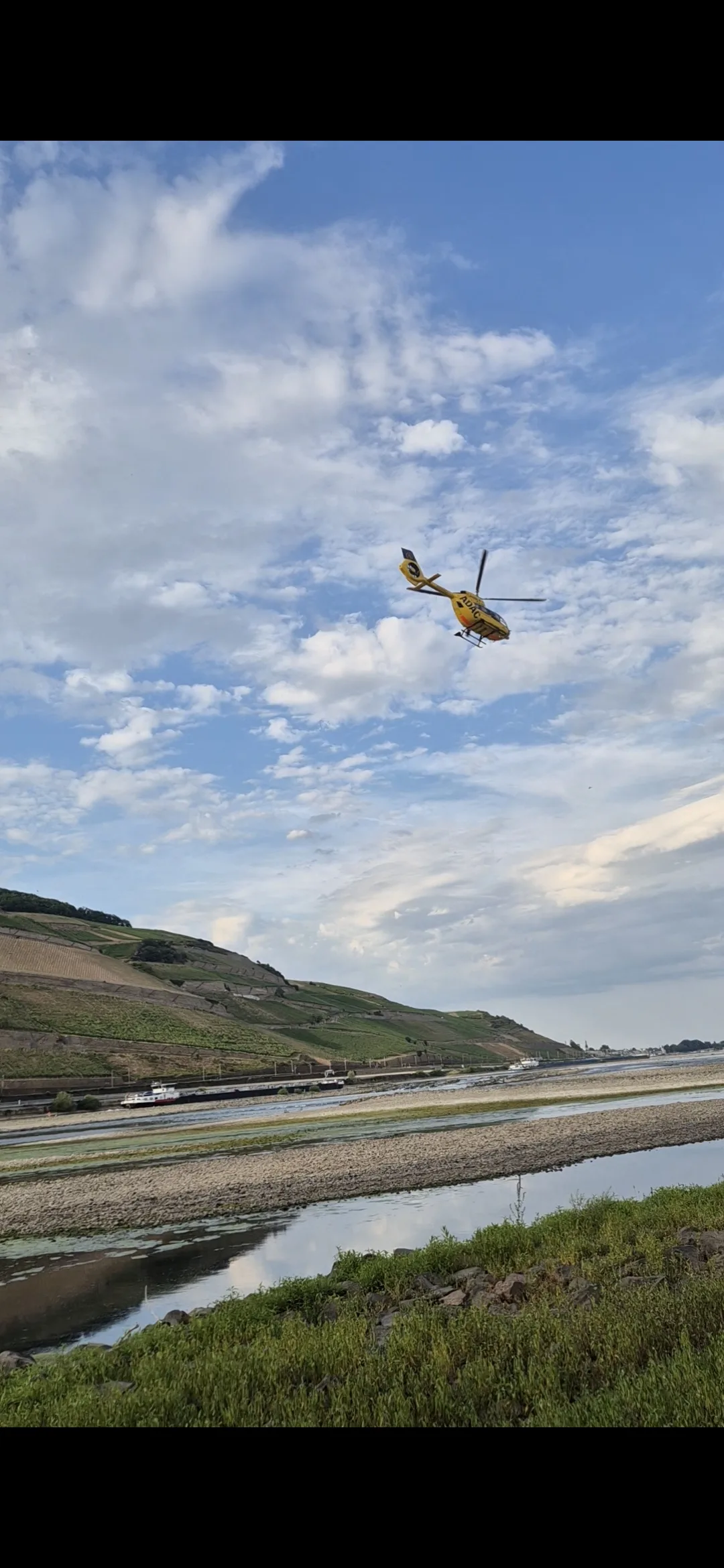 Ein gelber Rettungshubschrauber fliegt über einem Flusstal mit Weinbergen im Hintergrund. Der Himmel ist teils wolkig.