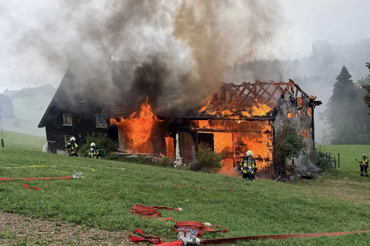 Ein großes Feuer hat ein Gebäude erfasst, dessen Dach bereits eingestürzt ist. Feuerwehrleute in Schutzanzügen versuchen, das Feuer zu löschen.