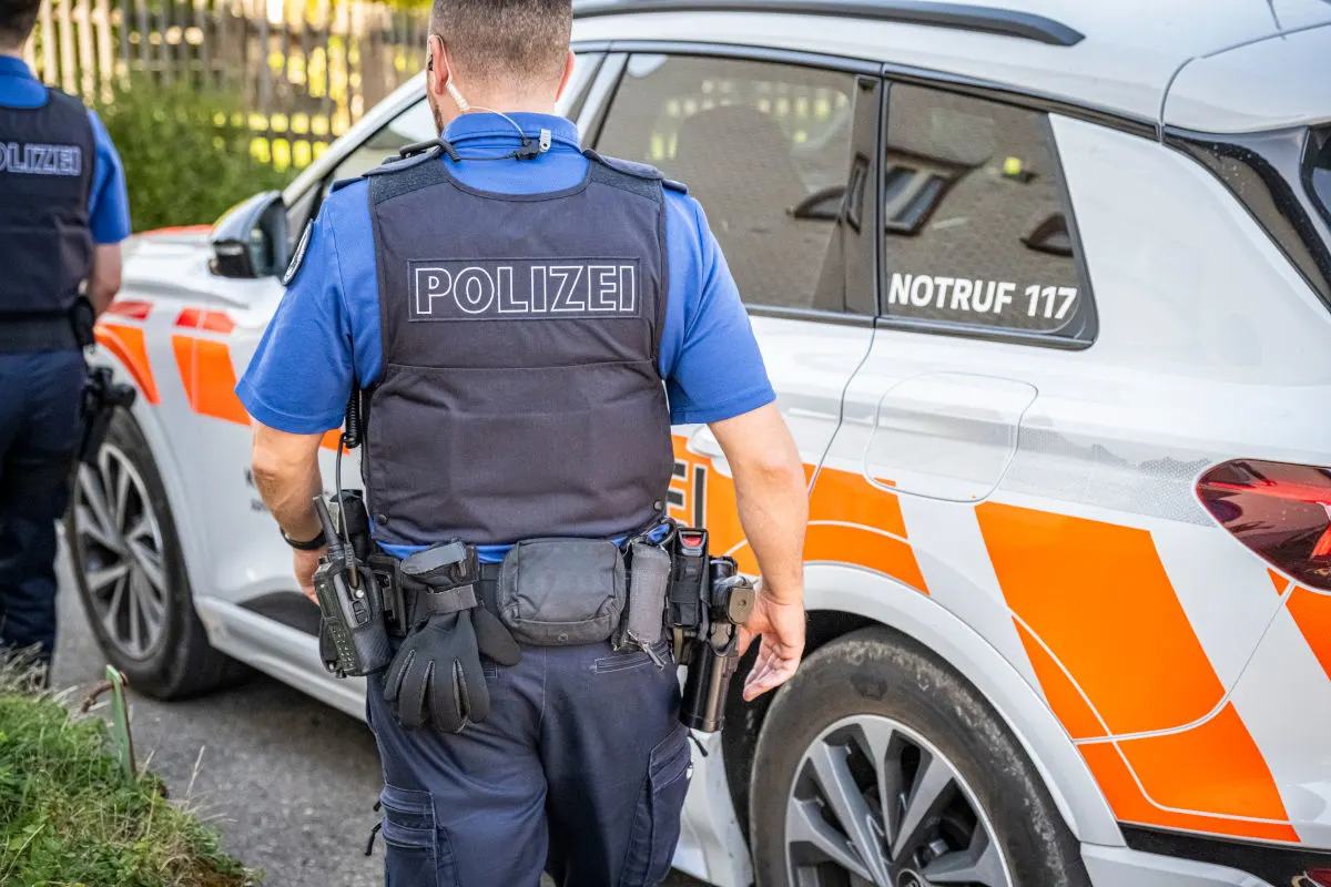 Ein Polizist steht neben einem weiß-orangefarbenen Polizeiauto mit der Aufschrift 'Polizei' und der Notrufnummer 117. Der Polizist trägt eine Weste mit 'Polizei' Aufschrift.