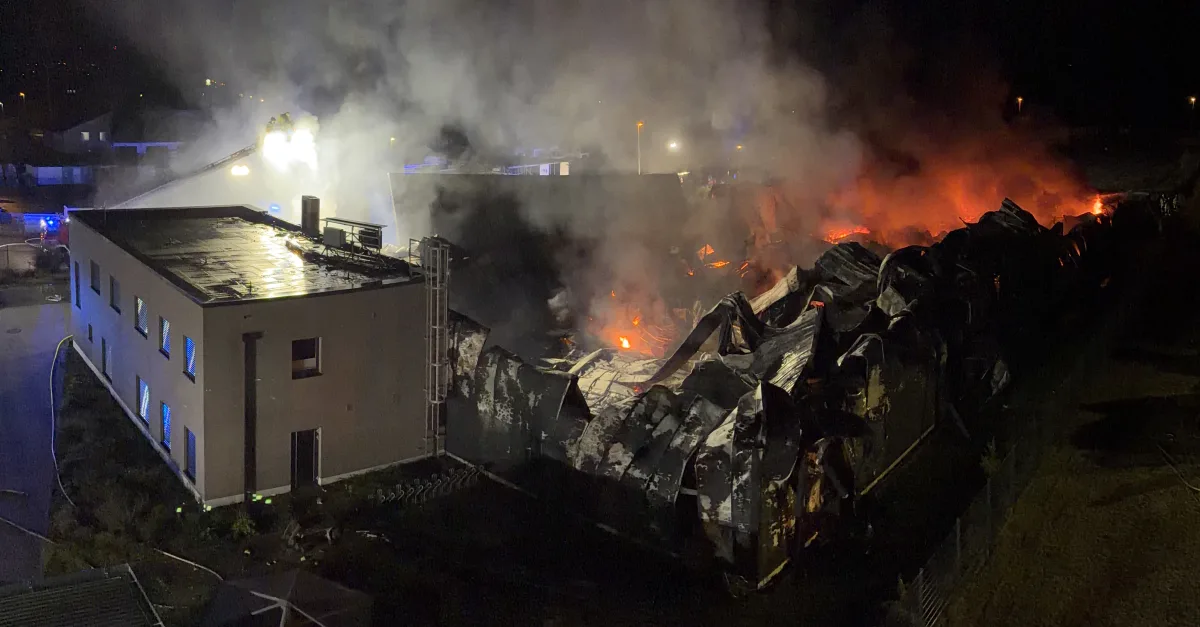 Ein Industriegebäude steht nachts in Flammen, während die Feuerwehr den Brand bekämpft. Dichte Rauchschwaden steigen in den Himmel.