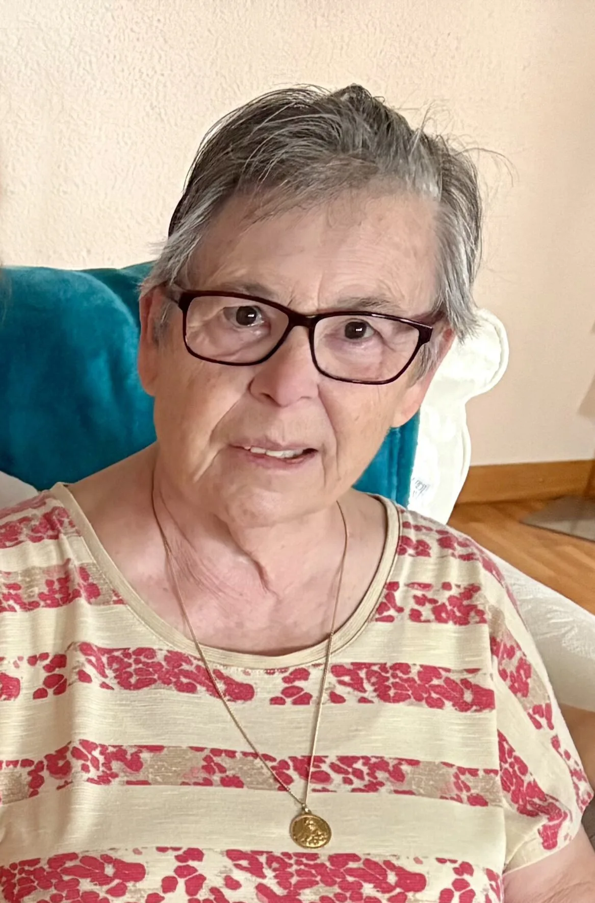 Das Bild zeigt eine ältere Person mit Brille, grauem Haar und einem gestreiften Oberteil. Sie sitzt auf einem Sofa.
