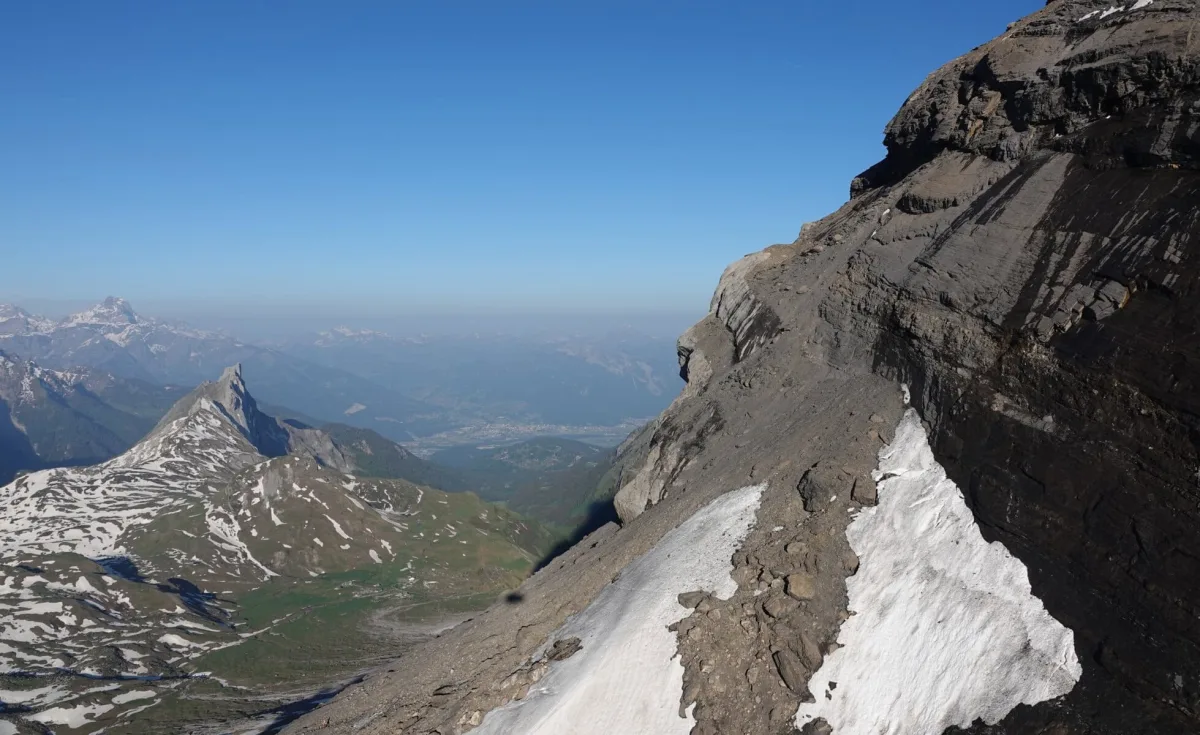 Eine steile Felswand mit Schneeresten liegt im Vordergrund. Im Hintergrund erstreckt sich eine bewaldete Berglandschaft.