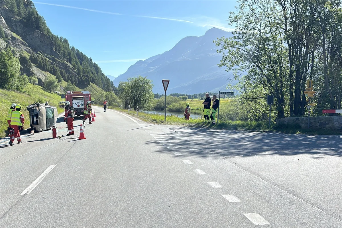 Ein Fahrzeug liegt auf der Seite auf einer Landstraße, umgeben von Rettungskräften in einer Berglandschaft bei klarem Wetter.