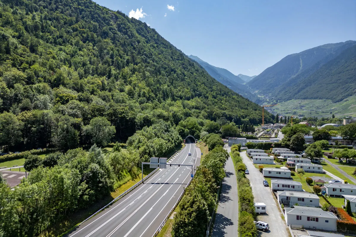 Das Bild zeigt eine Straße, die in einen Tunnel führt, umgeben von einer bewaldeten Berglandschaft an einem sonnigen Tag. Im Vordergrund sind Wohnwagen zu sehen.