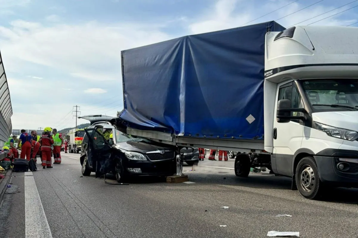 Auf einer Autobahn ist ein Pkw in die Seite eines Lkw-Anhängers geklemmt. Rettungskräfte sind vor Ort, einige knien neben dem Fahrzeug.