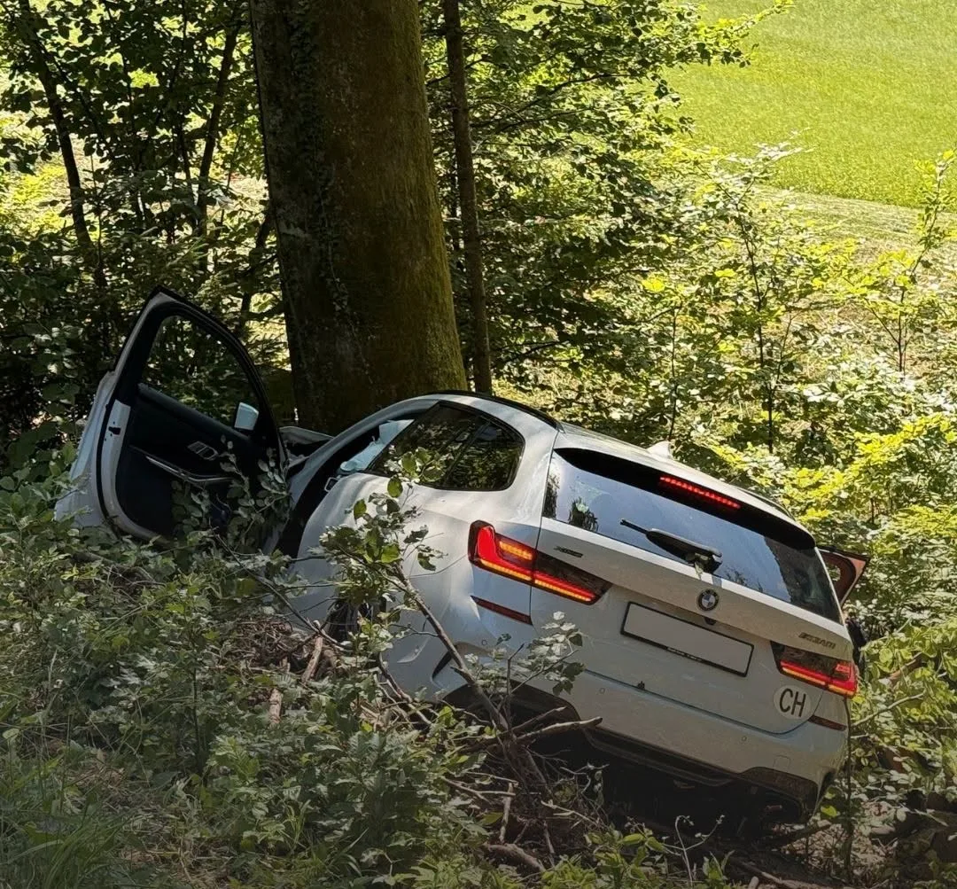 Ein weißer BMW, mit einem ovalen CH-Aufkleber, steht mit offener Tür in einem dichten Waldstück, umgeben von Bäumen und Vegetation.