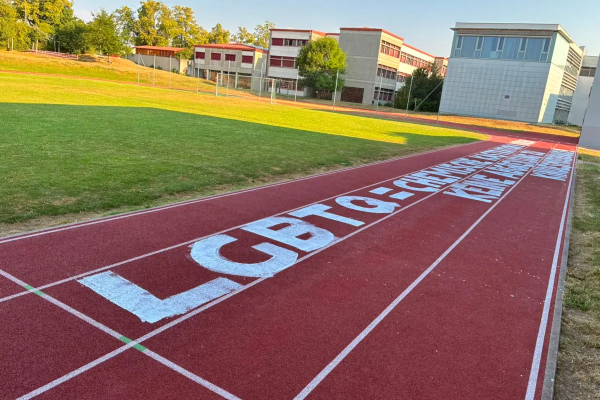 Die Laufbahn eines Sportplatzes zeigt weiße Buchstaben, die vor Schulgebäuden liegen. Der Platz ist von Bäumen umgeben und der Himmel ist klar.