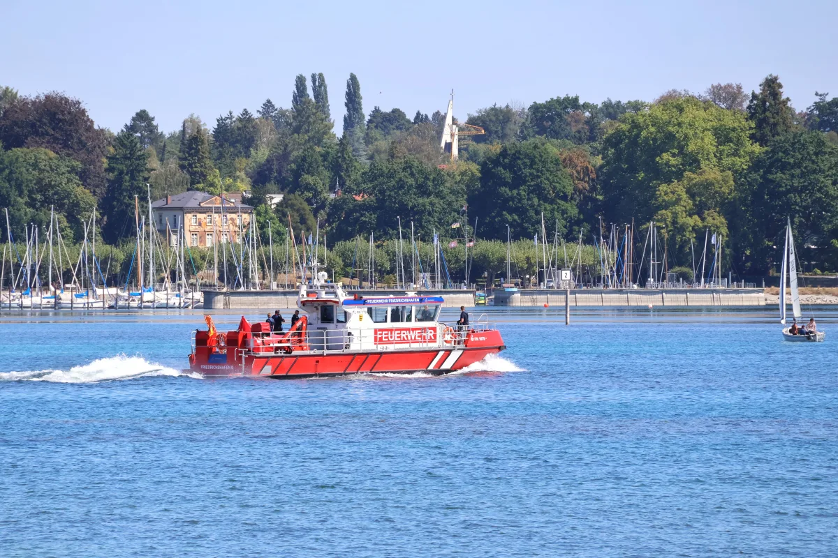Ein rotes Feuerwehrboot bewegt sich auf einem See vor mehreren Segelbooten und einer bewaldeten Uferlandschaft. Der Himmel ist klar.