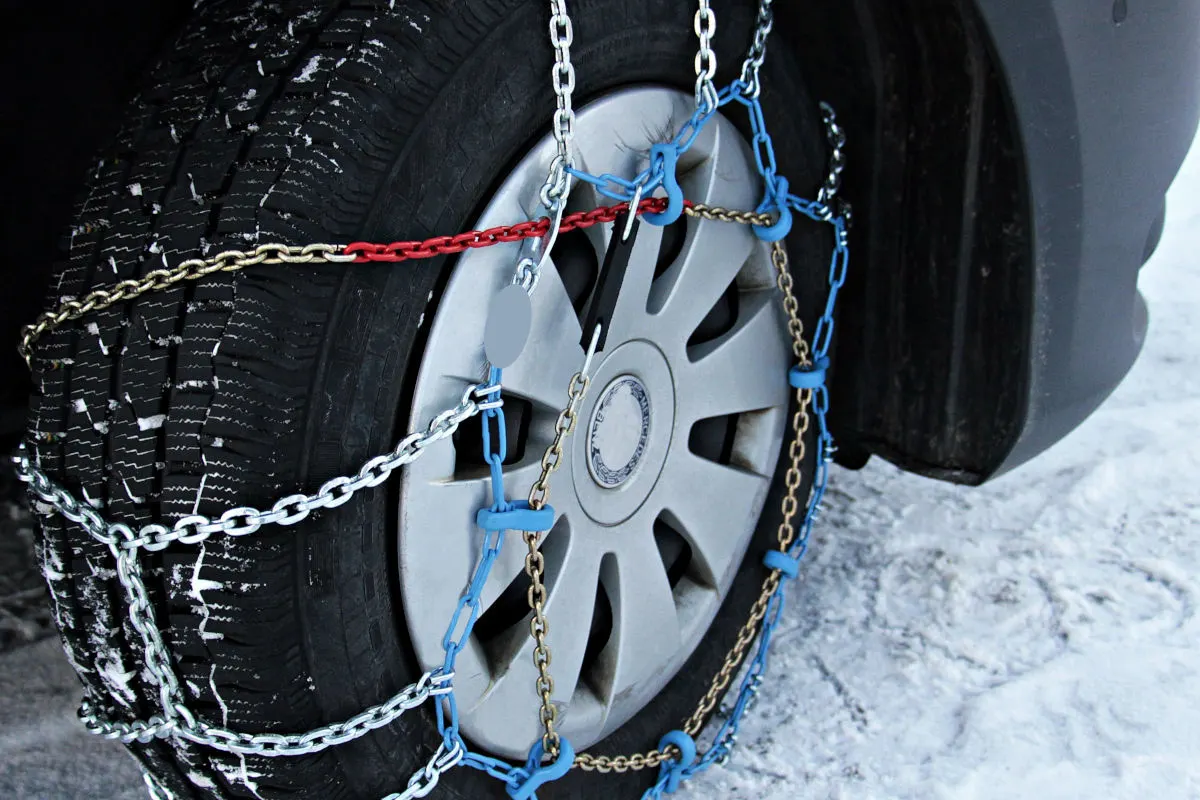 Ein Autoreifen ist mit Schneeketten versehen, die auf einer schneebedeckten Straße montiert sind. Die Ketten sind sorgfältig um den Reifen angebracht, um die Traktion bei winterlichen Bedingungen zu verbessern.