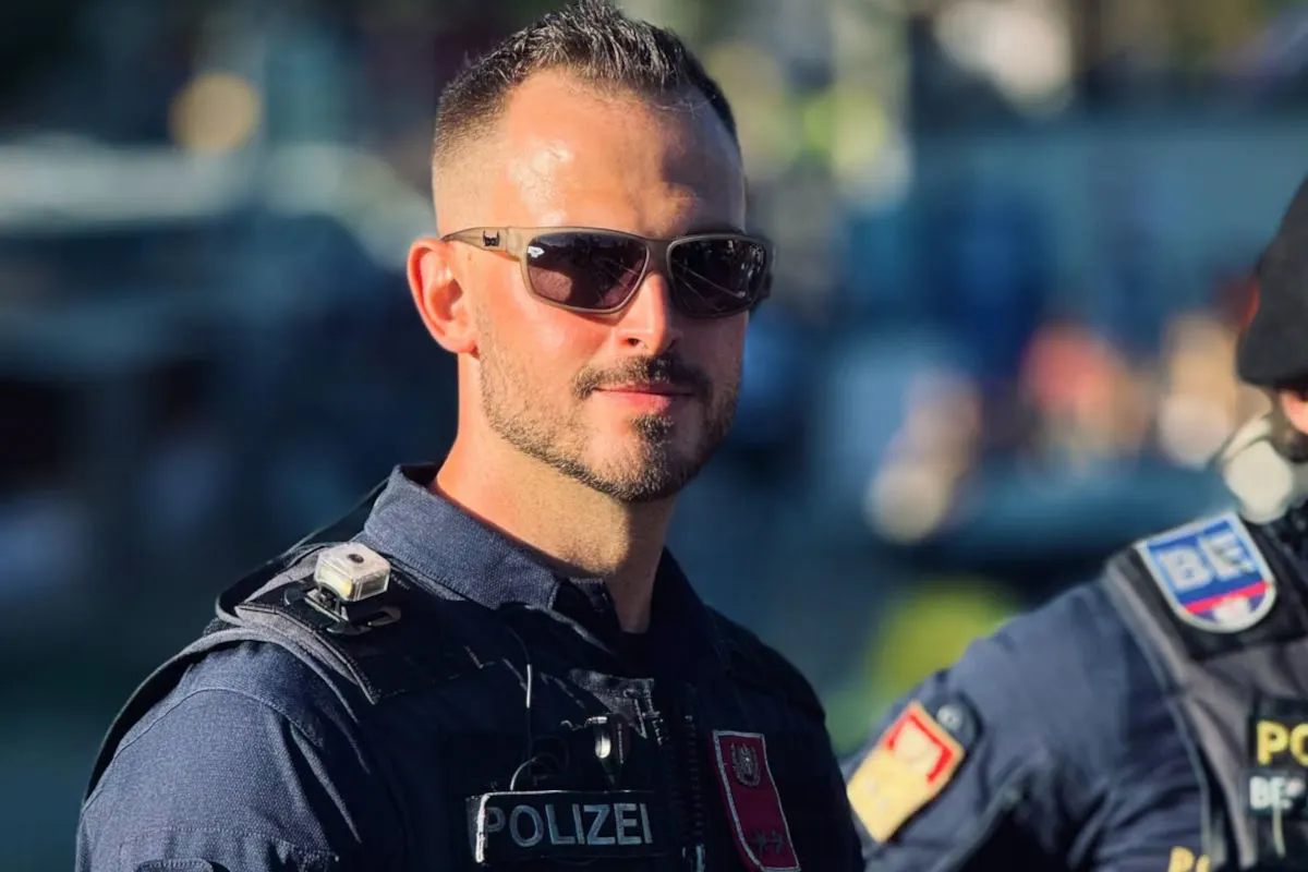 Ein Polizist mit Sonnenbrille trägt eine blaue Polizeiuniform und steht im Fokus, während der Hintergrund verschwommen ist. Auf seiner Weste sind Abzeichen sichtbar.