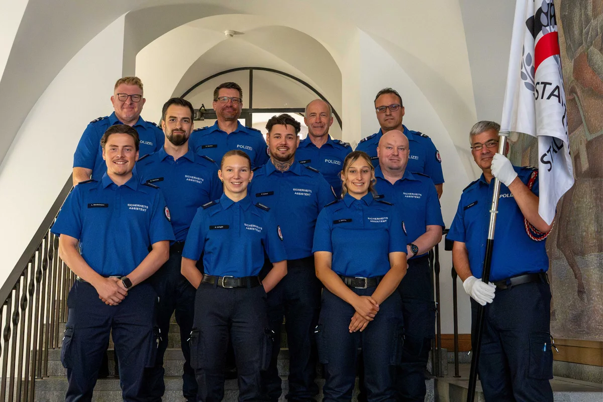 Eine Gruppe von Sicherheitsassistenten in blauen Uniformen steht lächelnd auf einer Treppe in einem Gebäude, neben ihnen hängt eine Fahne.