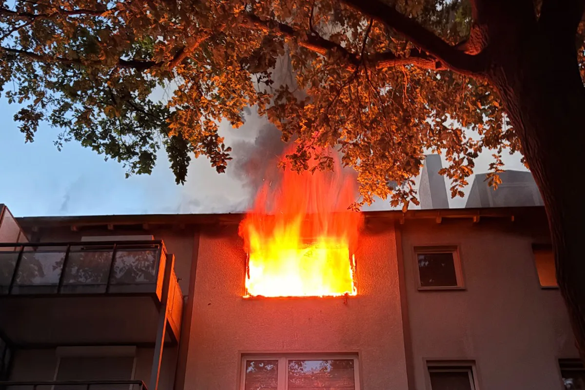 Leuchtende Flammen dringen aus einem Fenster im oberen Stockwerk eines Gebäudes hervor, umgeben von Rauch; ein Baum steht im Vordergrund.