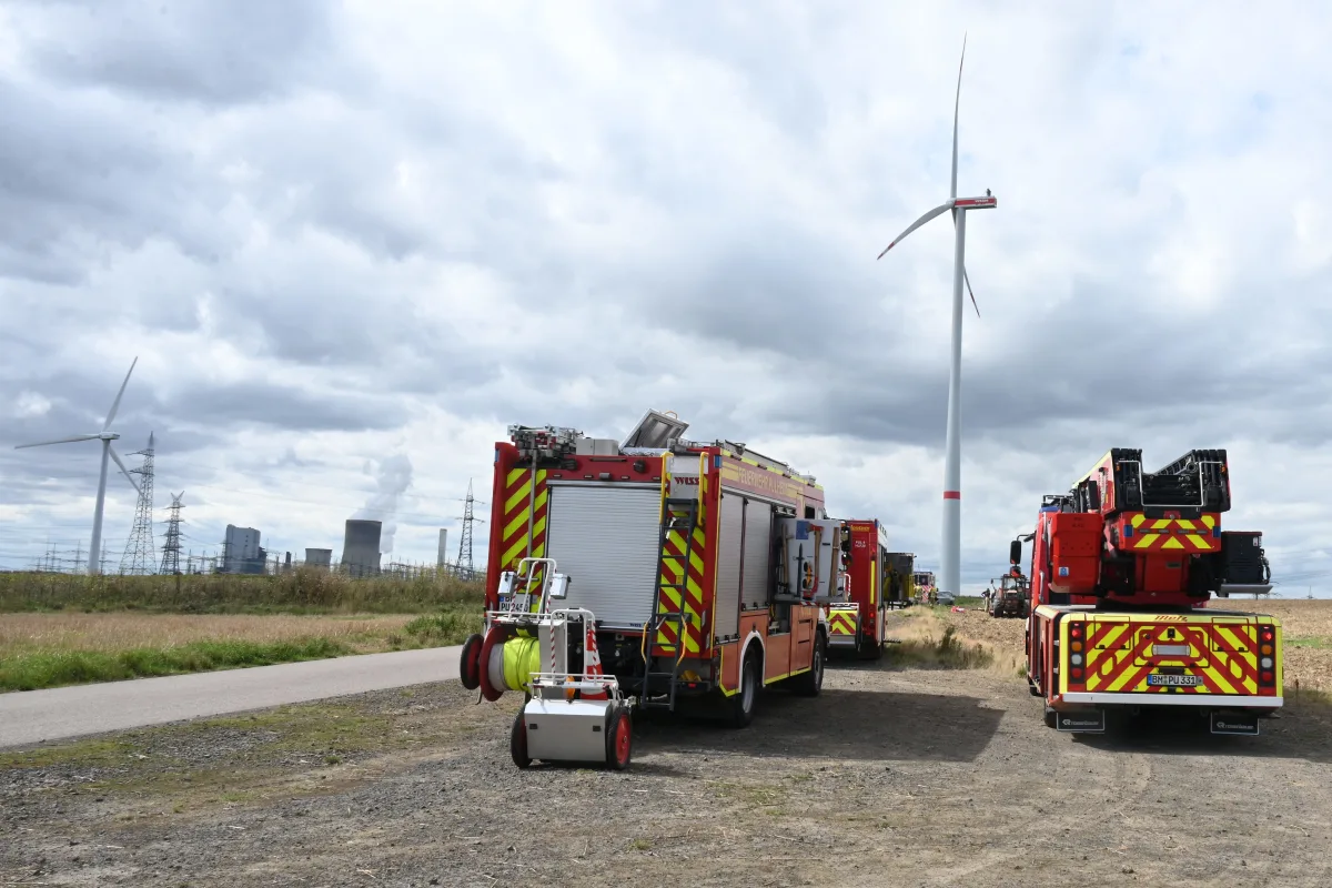 Mehrere Feuerwehrfahrzeuge sind in einem ländlichen Gebiet im Einsatz. Im Hintergrund sind Windkraftanlagen und ein Industriekomplex zu sehen. Der Himmel ist bewölkt.