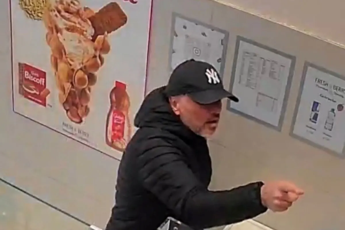 Ein Mann in dunkler Kleidung und Mütze steht in einem Geschäft und zeigt mit der Hand. Im Hintergrund sind Werbeschilder erkennbar.