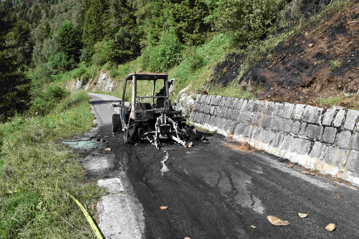 Das Bild zeigt ein völlig ausgebranntes Fahrzeug auf einer kurvigen Straße in einer bergigen Gegend. Die Umgebung ist grün, mit Bäumen und einer Steinmauer entlang der Straße.