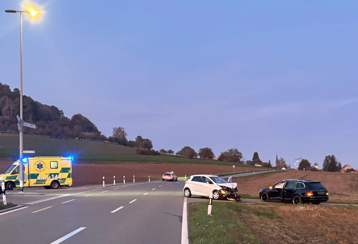 Eine ländliche Straße zeigt zwei beschädigte Autos nach einem Unfall. Ein Ambulanzfahrzeug steht mit eingeschaltetem Blaulicht am Straßenrand.