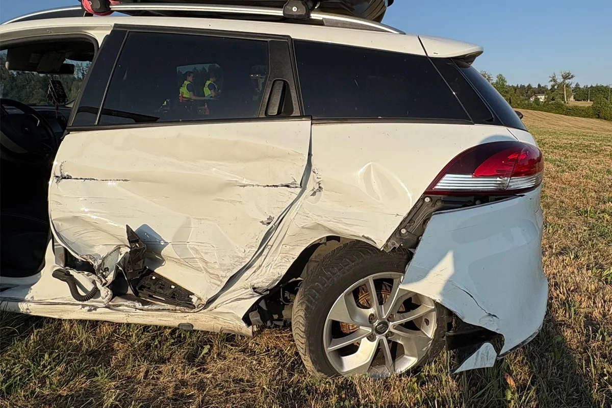 Ein weißes Auto mit schwer beschädigtem Heck steht auf einem Feld. Im Inneren sitzen Menschen mit gelben Warnwesten.