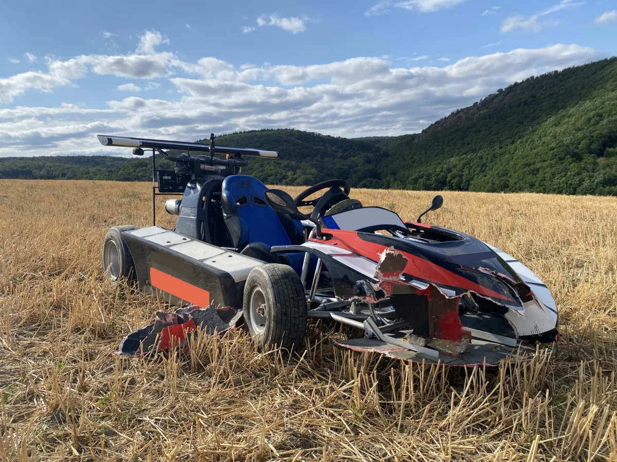 Ein stark beschädigter Rennwagen steht auf einem abgeernteten Feld. Der Himmel ist klar mit wenigen Wolken.