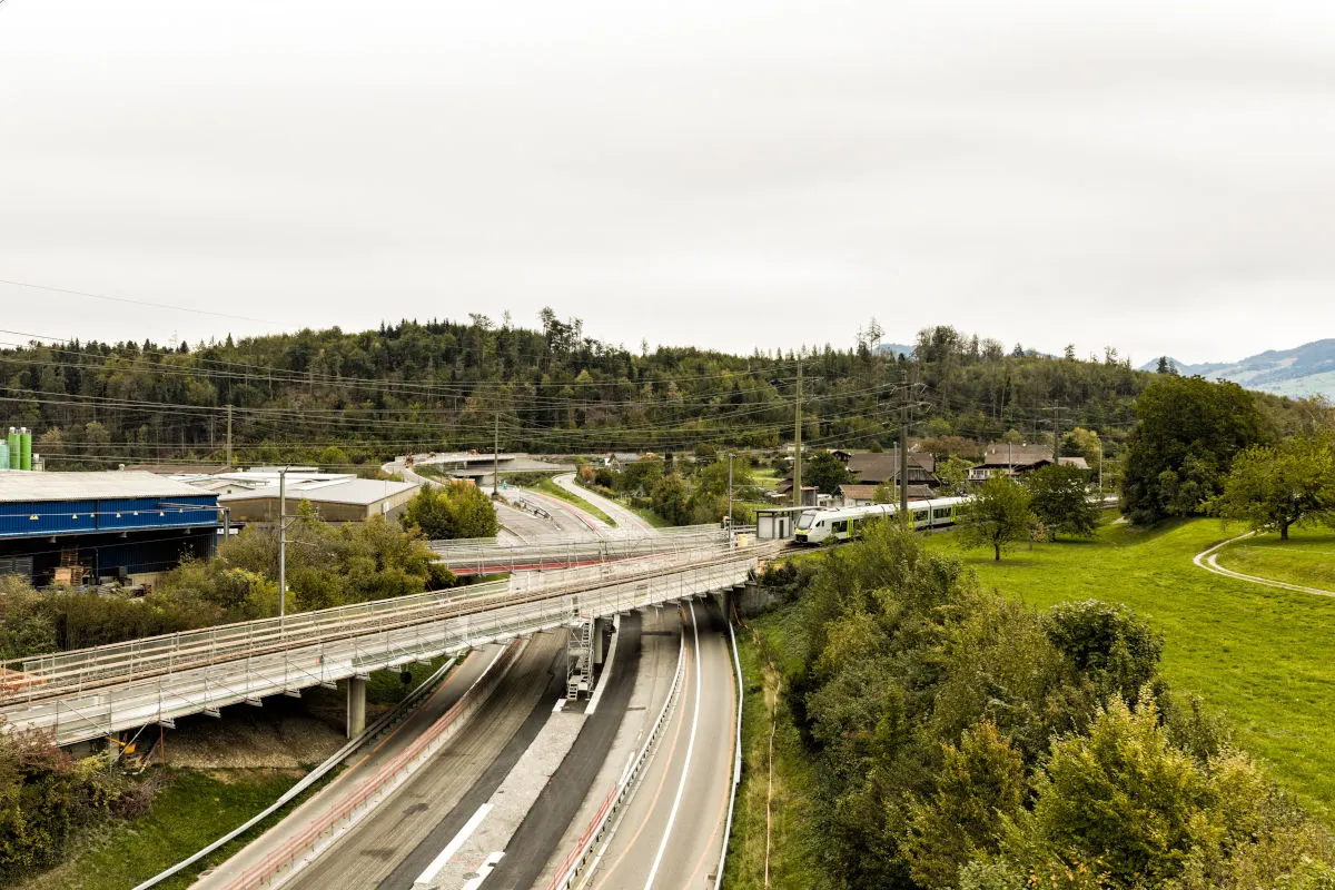 Eine moderne Brücke führt über eine mehrspurige Autobahn, im Hintergrund fährt ein Zug durch eine grüne Landschaft.