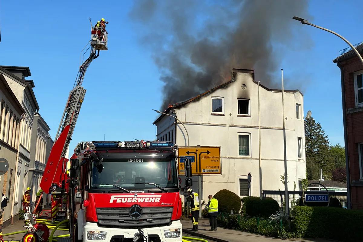 Ein Feuerwehrfahrzeug mit einem Leiteraufbau steht vor einem Gebäude, aus dessen Dach schwarze Rauchwolken aufsteigen. Feuerwehrleute sind im Einsatz.