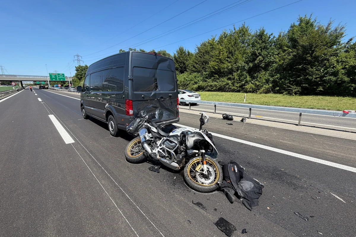 Ein Motorrad steht beschädigt vor einem dunklen Transporter auf einer Autobahn, daneben liegen Trümmerteile. Im Hintergrund sind weitere Fahrzeuge und eine grüne Landschaft sichtbar.