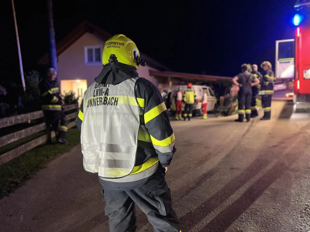 Ein Feuerwehrmann mit gelbem Helm und reflektierender Weste steht auf einer Straße. Im Hintergrund sind weitere Einsatzkräfte und ein Feuerwehrauto zu sehen.