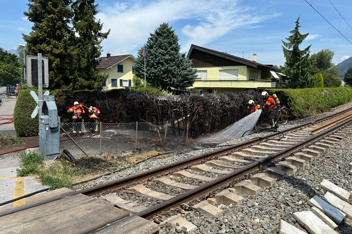 Feuerwehrleute in Schutzbekleidung löschen einen Heckenbrand neben Bahngleisen in einem Wohngebiet. Zwei Häuser sind im Hintergrund erkennbar. Der Einsatz findet bei klarem Himmel statt.