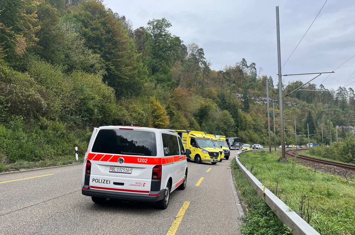 Auf einer Landstraße nahe Basel stehen ein Polizeifahrzeug der Kantonspolizei Basel-Landschaft und mehrere Rettungswagen. Üppige Vegetation und Bahngleise sind neben der Straße sichtbar.